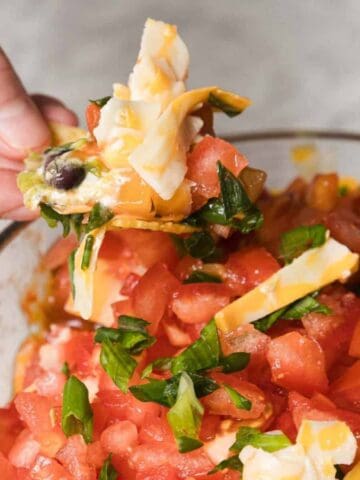 A person holds a chip topped with diced tomatoes, green onions, cheese, and olives, over a bowl filled with the same ingredients. The background is a light-colored countertop.