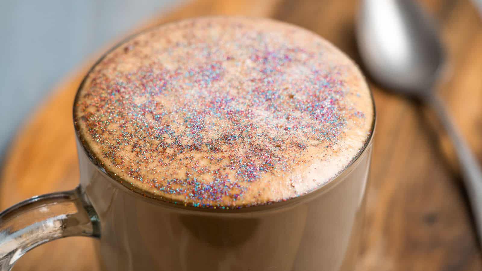 A glass mug filled with coffee or hot chocolate is topped with a frothy layer sprinkled with multicolored glitter. A spoon rests on a wooden surface in the background.