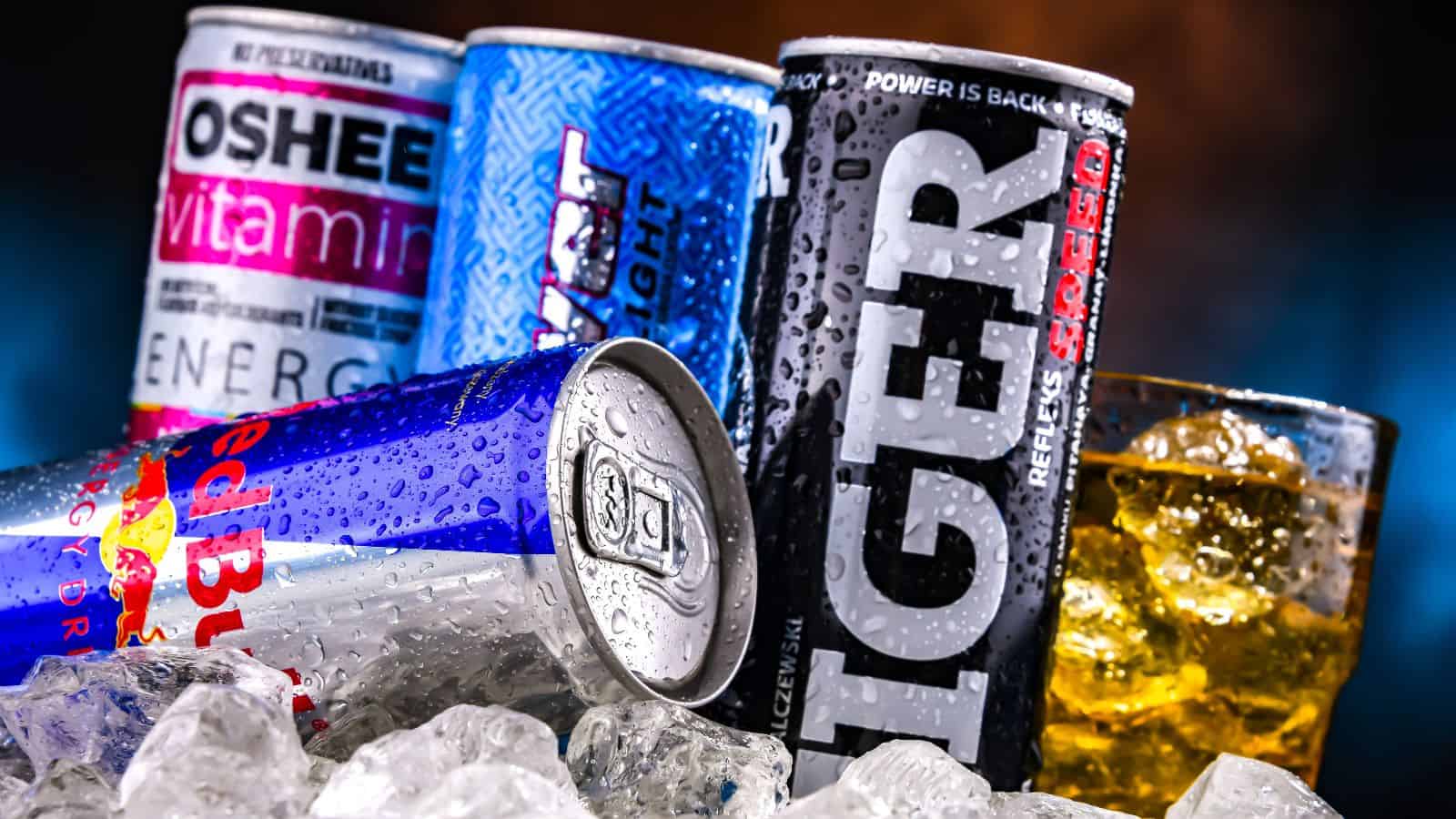 Cans of various energy drinks, including Oshee, Blue Motion, Tiger, and Red Bull, are placed on ice. A glass filled with an amber liquid and ice cubes is beside the cans. Water droplets are present on the cans, indicating coldness.