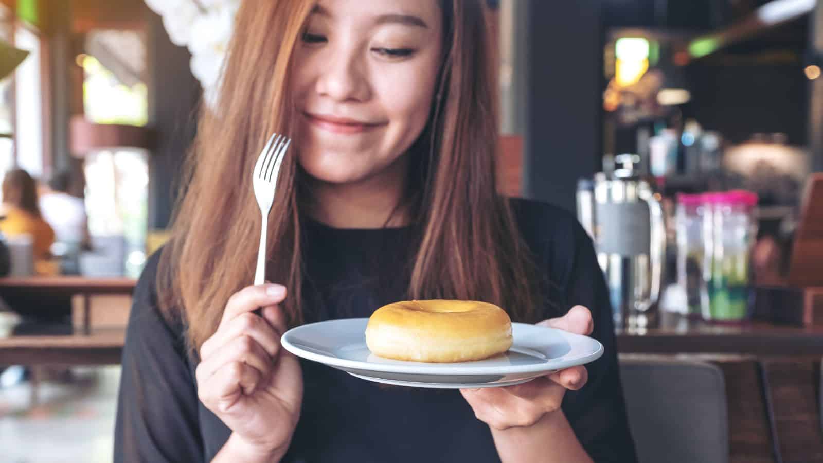 A woman with long hair smiles while holding a white plate with a yellow donut. She is holding a fork in her other hand. The background shows a blurred indoor cafe setting.