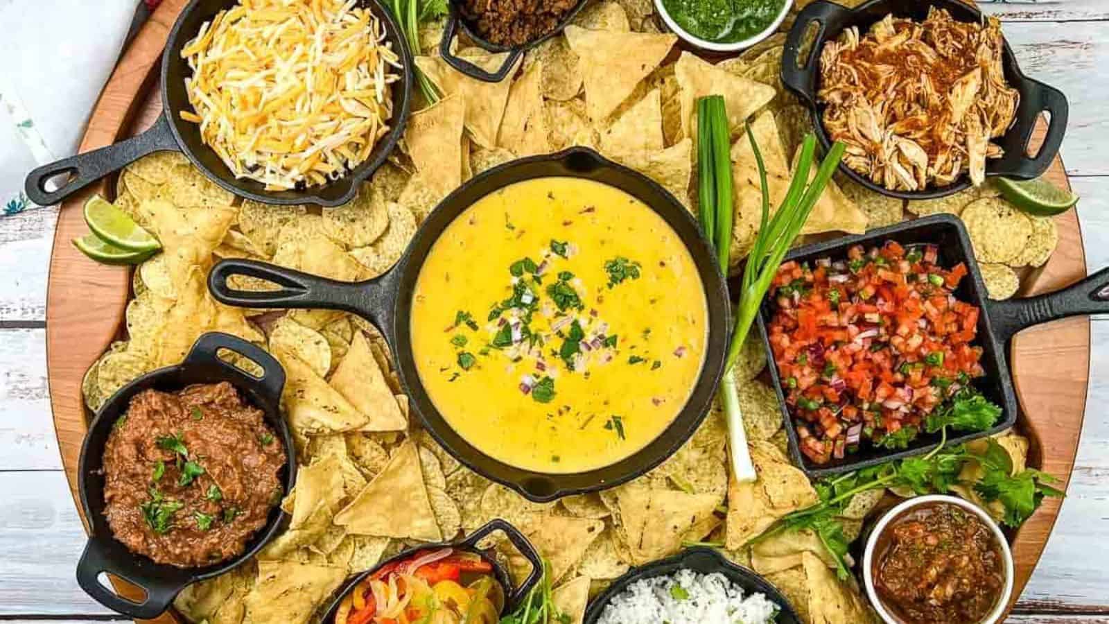 A platter with a large bowl of cheese dip in the center, surrounded by tortilla chips. Around are bowls containing shredded cheese, salsa, guacamole, shredded chicken, ground beef, beans, mixed vegetables, and toppings like green onions and cilantro.