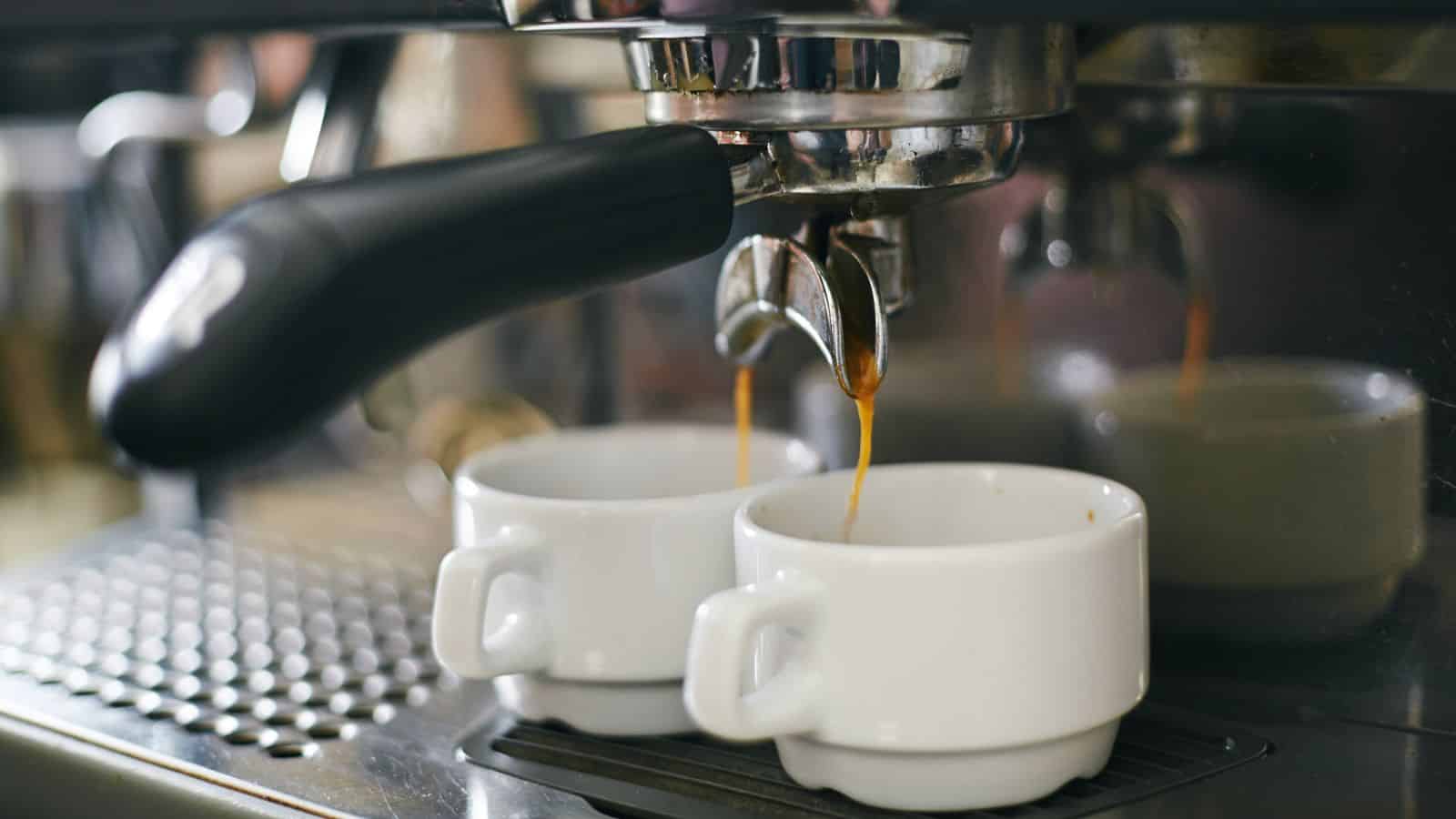 A close-up of an espresso machine brewing coffee into two white ceramic cups. The liquid is visibly pouring from the spouts, and the background shows the metallic surface of the machine.