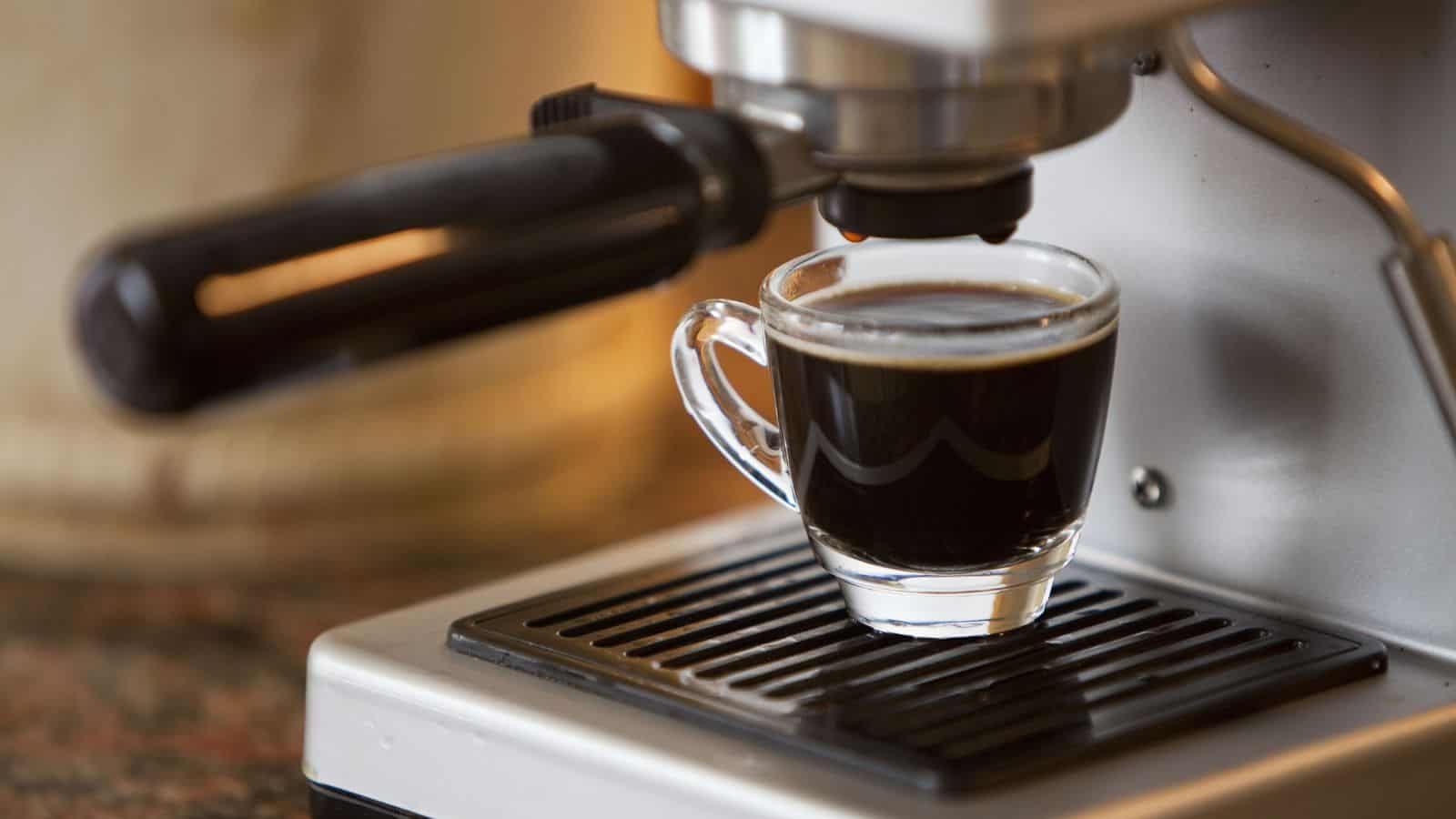 A close-up image of an espresso machine dispensing dark coffee into a clear glass cup on a metal tray. The machine is silver and black with a visible handle, set against a blurred background.
