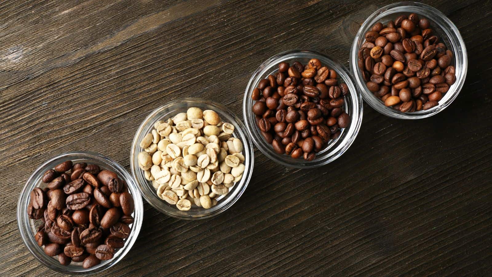 Four glass bowls on a wooden surface, each containing different types of coffee beans: one with dark roasted beans, one with unroasted green beans, and two with medium roasted beans.