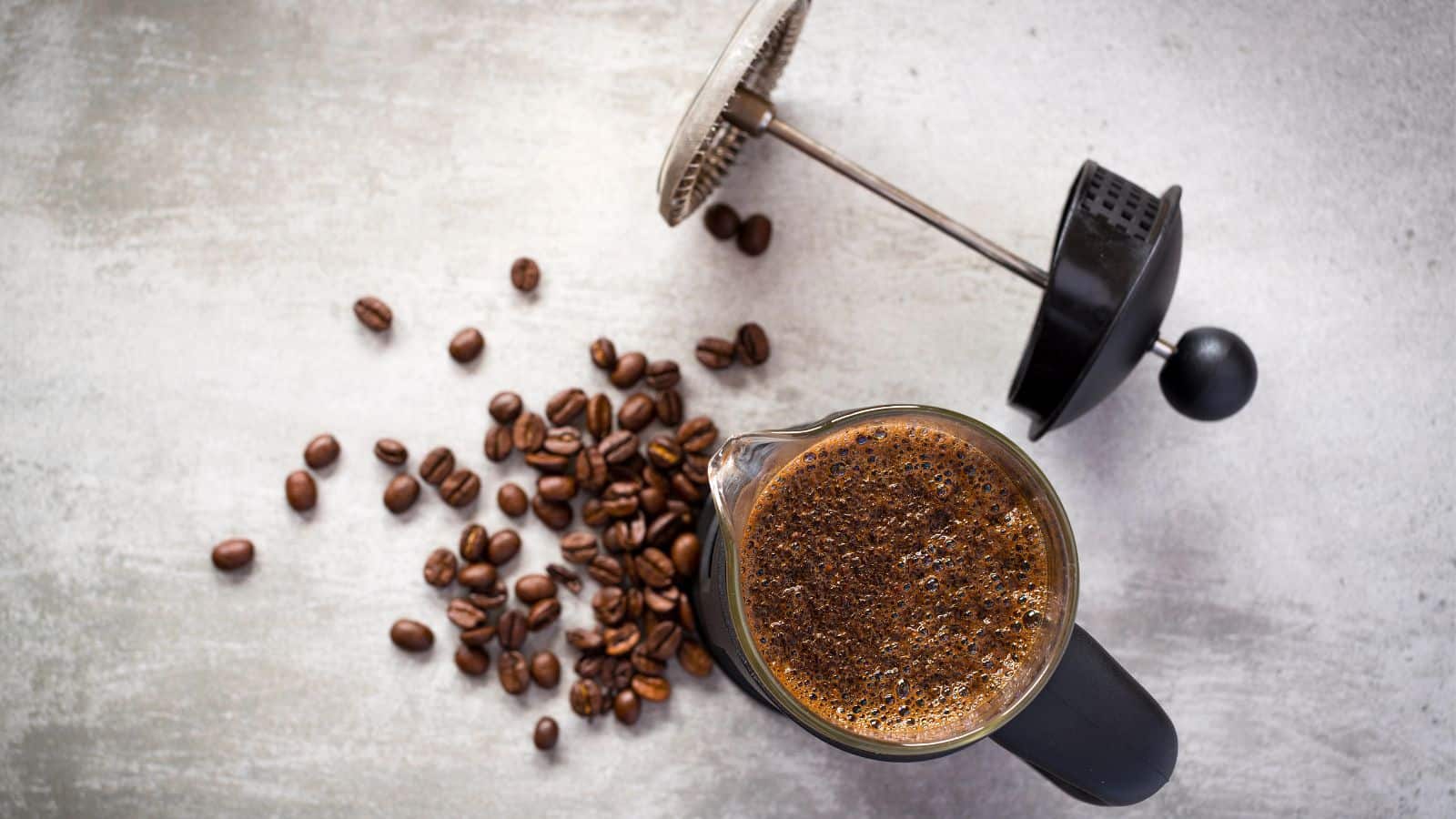 A French press with freshly brewed coffee and scattered coffee beans on a textured surface. The lid of the press is off to the side, revealing the coffee's rich, foamy top.