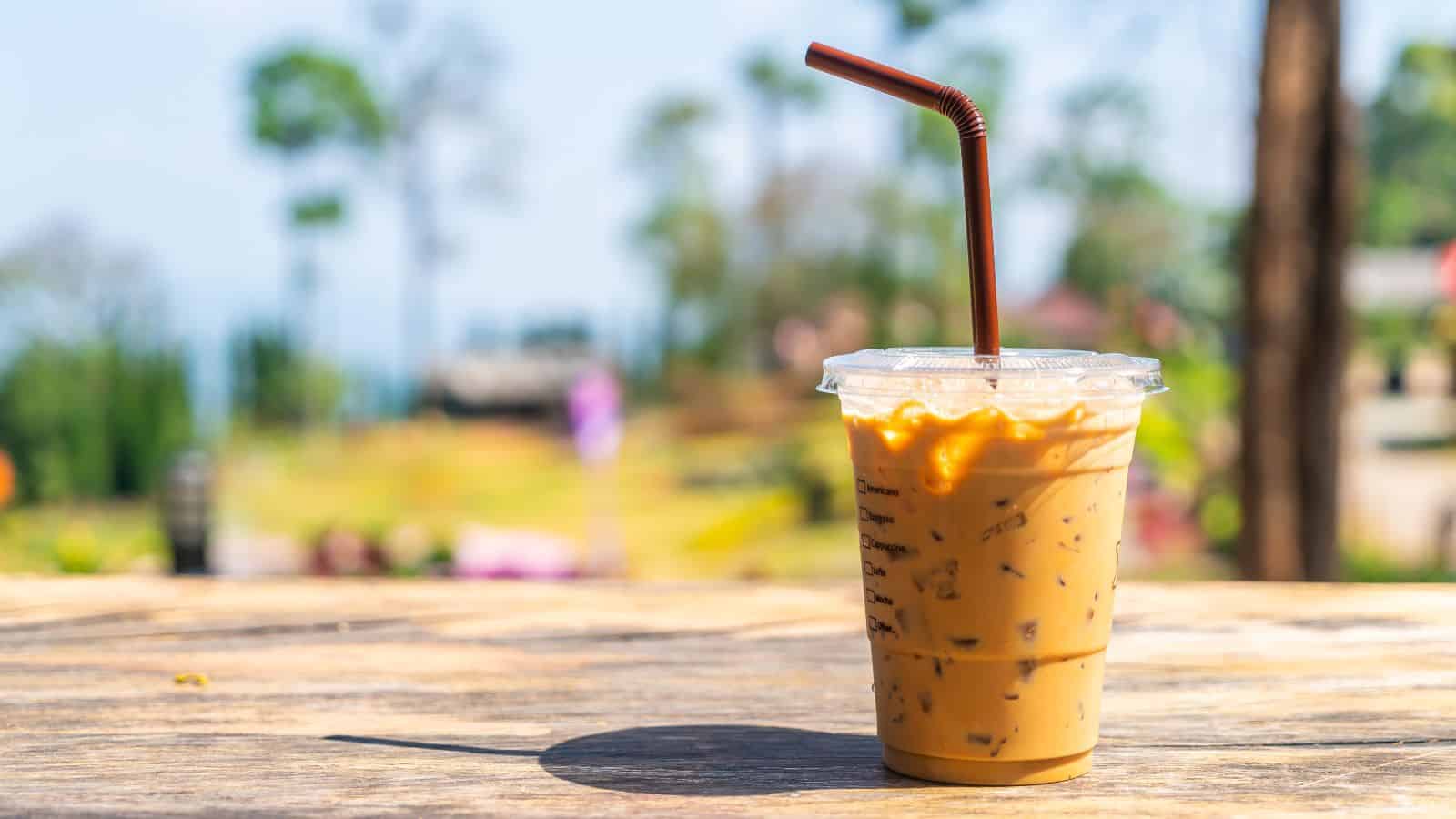 A plastic cup of iced coffee with a brown straw sits on a wooden table. The background is blurred with greenery and trees, suggesting an outdoor setting.