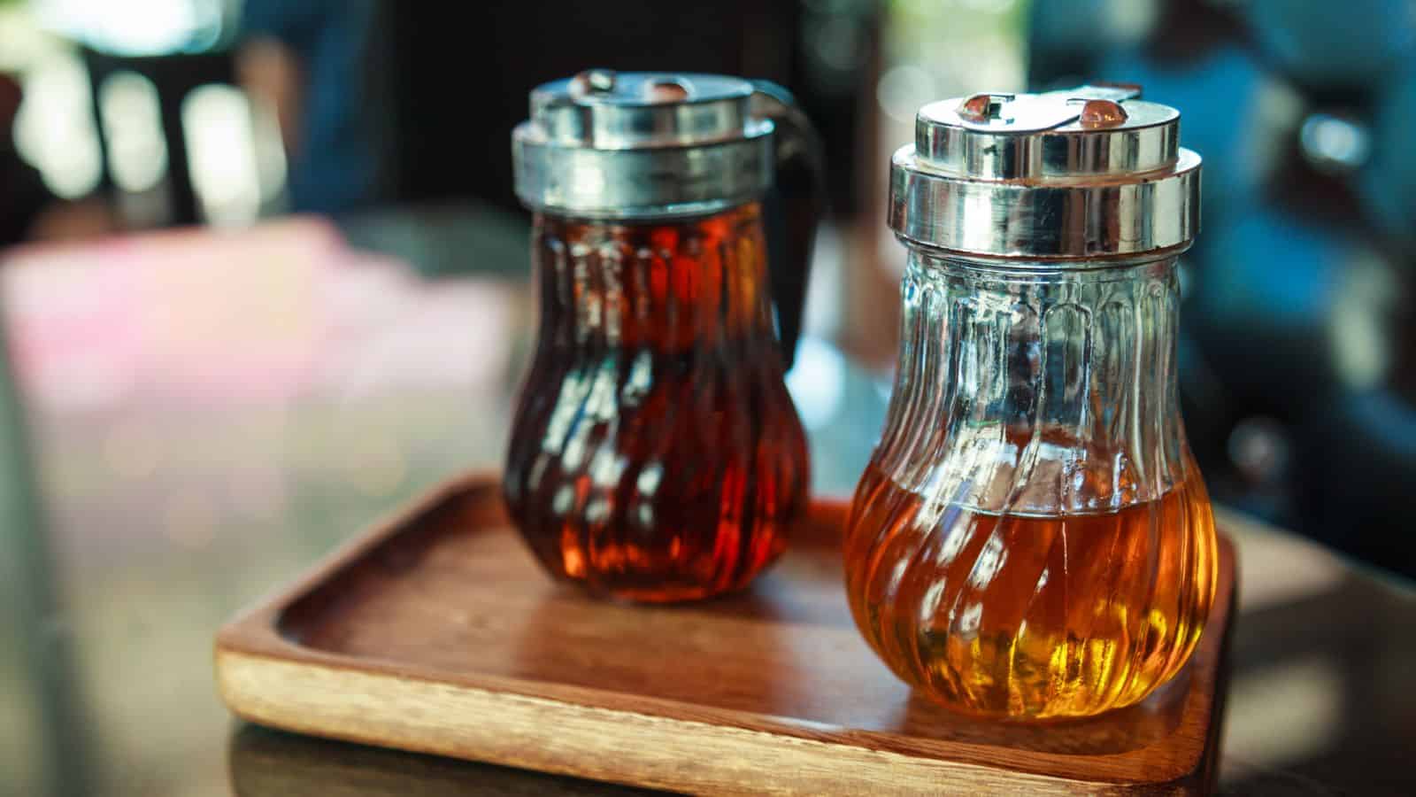 Two glass syrup dispensers with stainless steel lids sit on a wooden tray. One is filled with a dark liquid, the other with a lighter amber liquid. The background is blurred, suggesting an indoor setting.