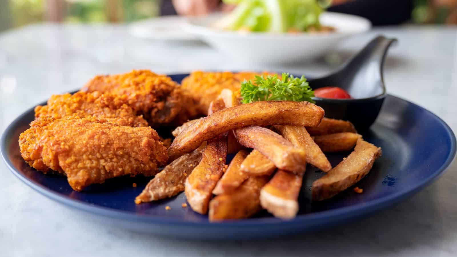 A blue plate with crispy fried chicken wings and seasoned fries is shown. A small black container with a cherry tomato and a sprig of parsley accompanies the dish on a marble table.