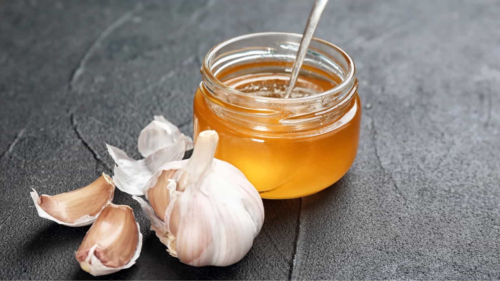 A jar of honey with a spoon sits on a dark surface. Next to it are several cloves of garlic, some peeled and some unpeeled.