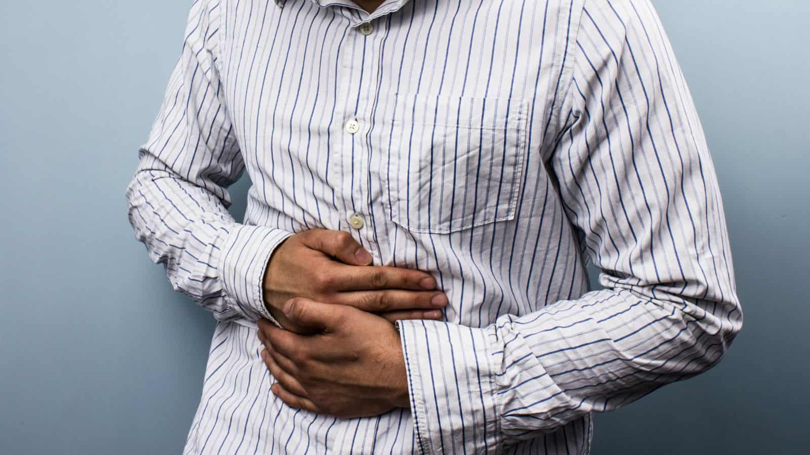 A person in a striped shirt holds their stomach with both hands, suggesting discomfort or pain. The individual's face is not visible, and they are standing against a plain, light-colored background.