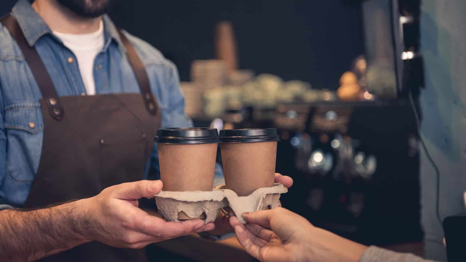 A barista wearing an apron hands two cardboard coffee cups in a holder to a customer. The setting appears to be a coffee shop, with blurred backgrounds of equipment and stacked cups.