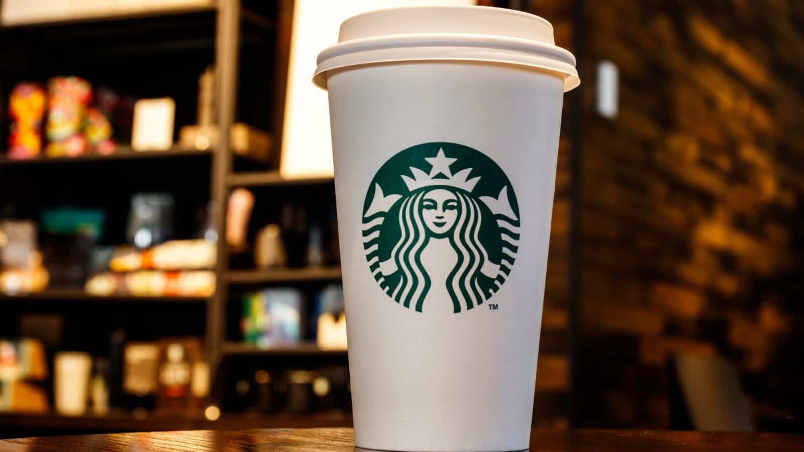 A white Starbucks coffee cup with a lid features the green Starbucks logo. The cup is on a wooden surface, with a blurred background of shelves and a brick wall, suggestive of a caf&eacute; setting.