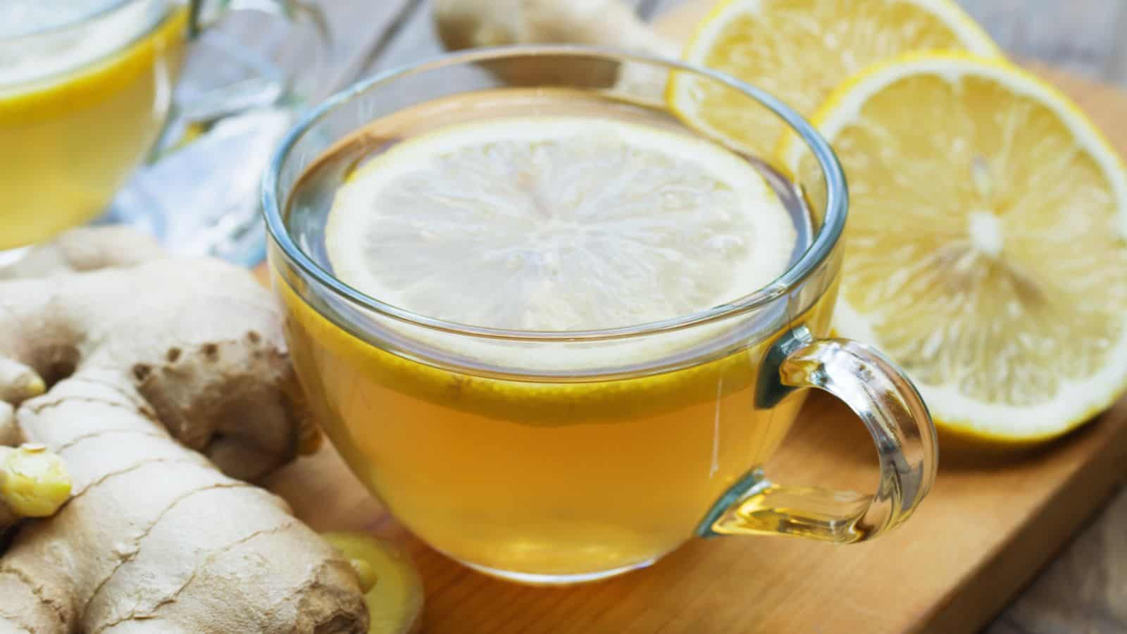 A clear glass cup of ginger tea with a slice of lemon on top rests on a wooden surface. Fresh ginger root and lemon slices are visible in the background, enhancing the tea's presentation.