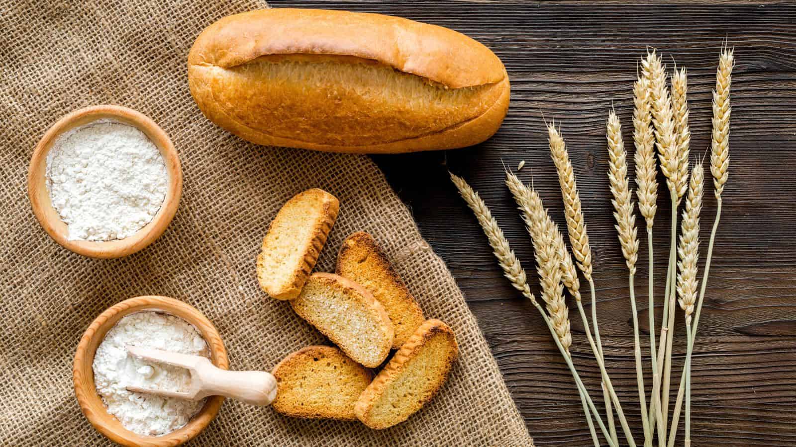 Loaf of bread and slices on burlap with wheat stalks. Two wooden bowls with flour and a small scoop are nearby. The scene is set on a dark wooden table.