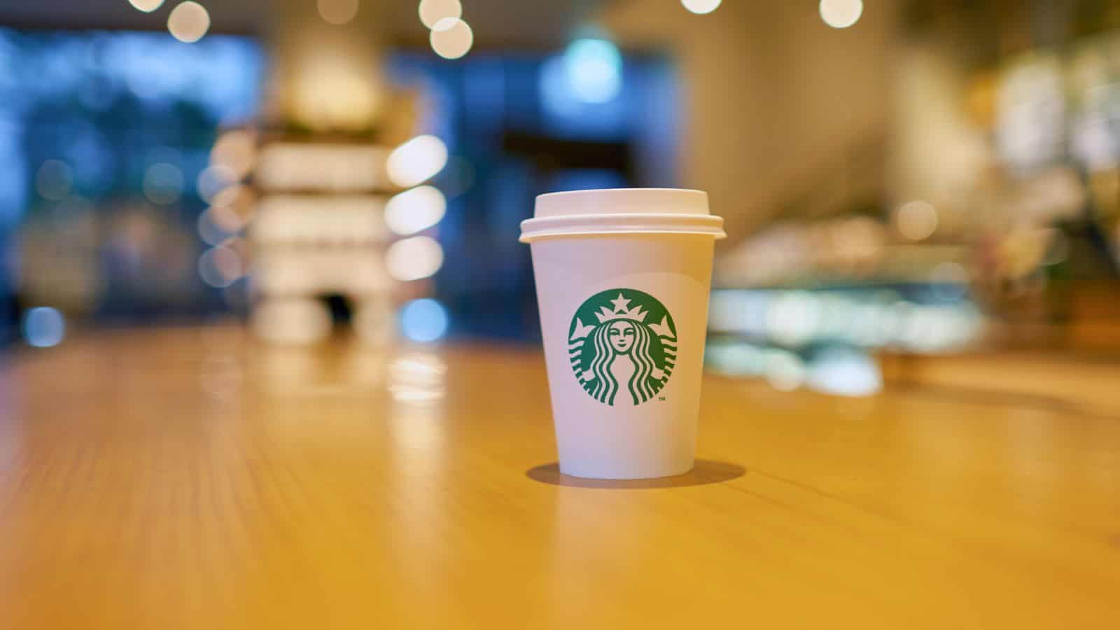 A white disposable cup with a green logo is placed on a wooden table. The background is softly blurred, indicating an indoor cafe setting with lights and shelves.