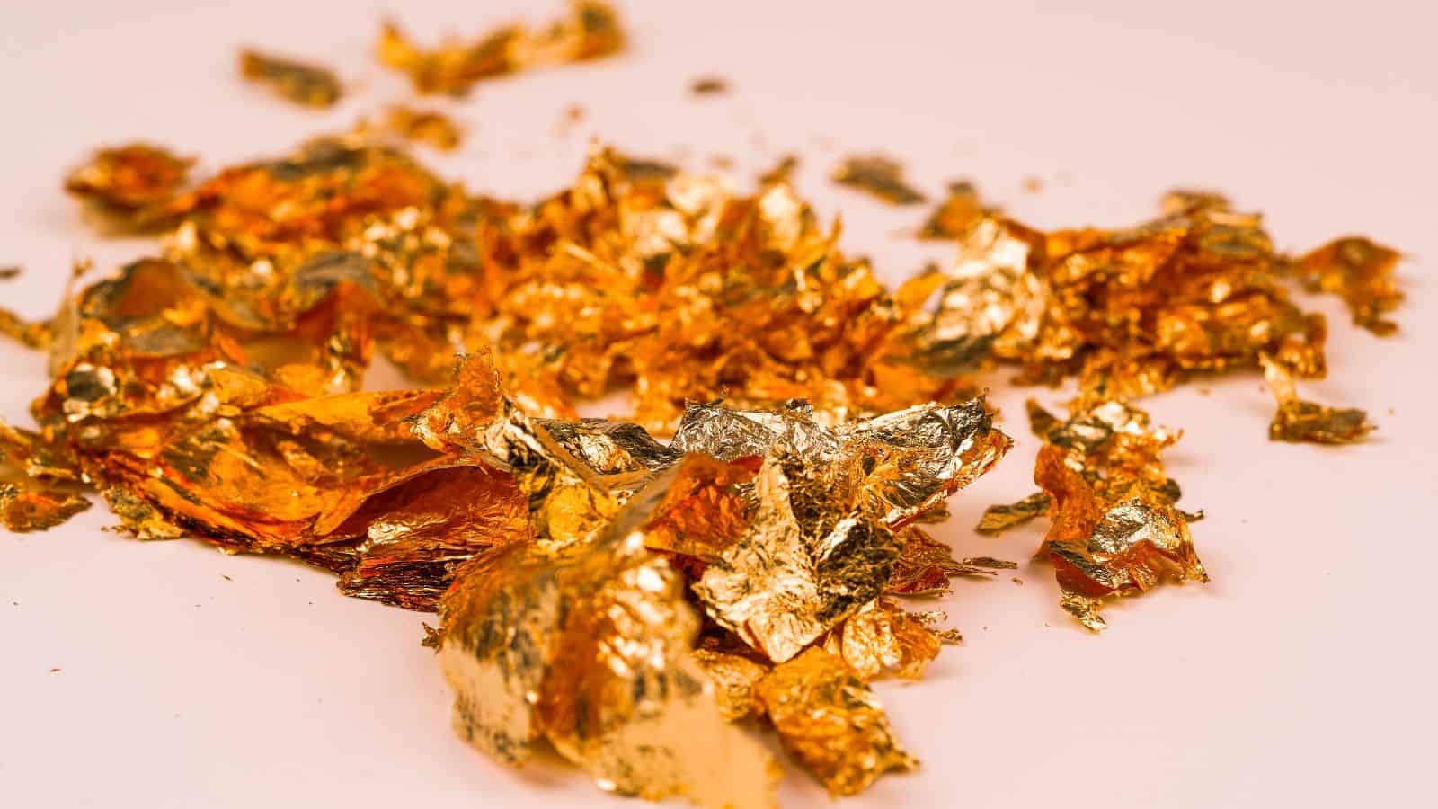Pieces of gold leaf scattered on a white surface. The gold leaf is crumpled and unevenly spread, displaying a shiny, metallic texture with various reflections.