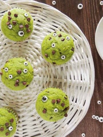 A basket holds seven green muffins decorated with small edible eyes and chocolate chips, placed on a wooden surface. To the right, one muffin sits on a white plate. Several edible eyes are scattered around the muffins.