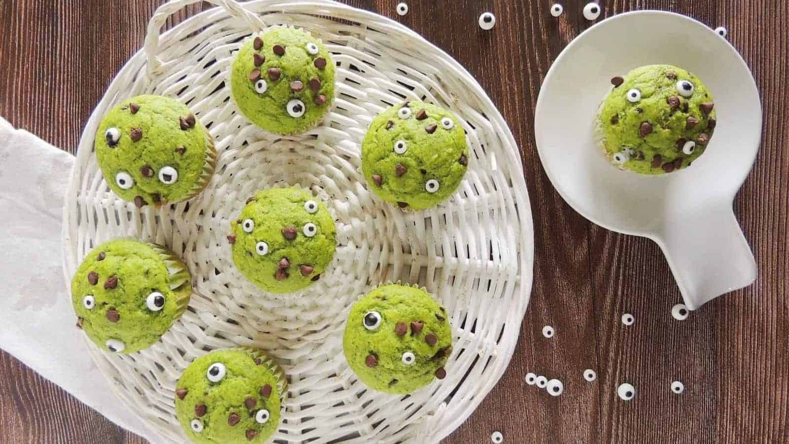 A basket holds seven green muffins decorated with small edible eyes and chocolate chips, placed on a wooden surface. To the right, one muffin sits on a white plate. Several edible eyes are scattered around the muffins.