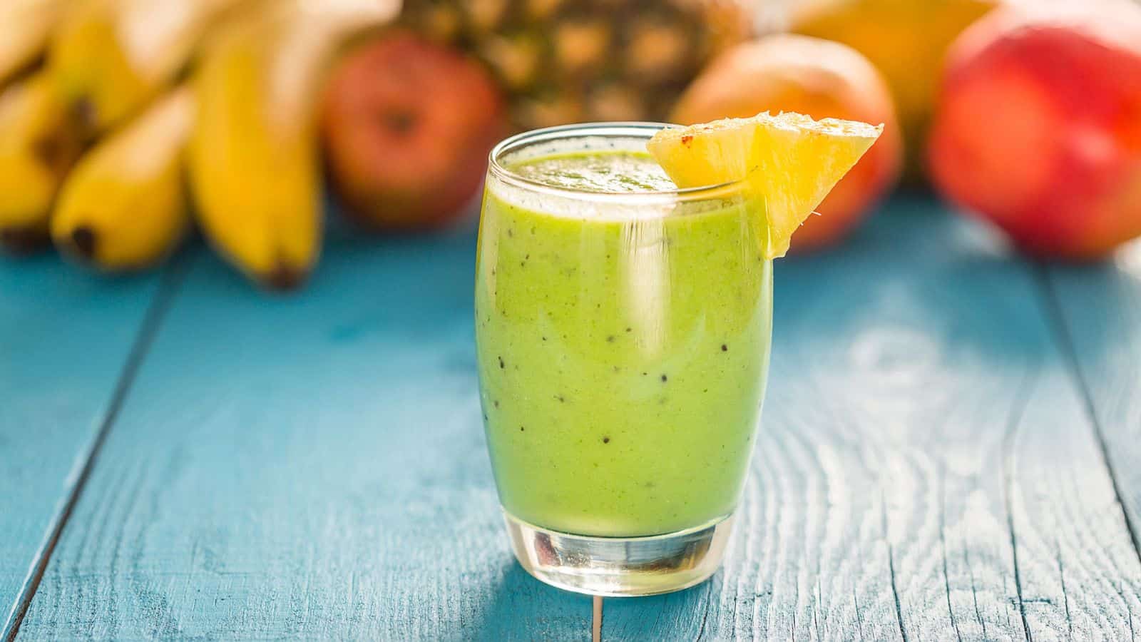 A glass of green smoothie on a blue wooden table, garnished with a pineapple slice. In the background, there are blurred fruits including bananas, apples, and a pineapple.
