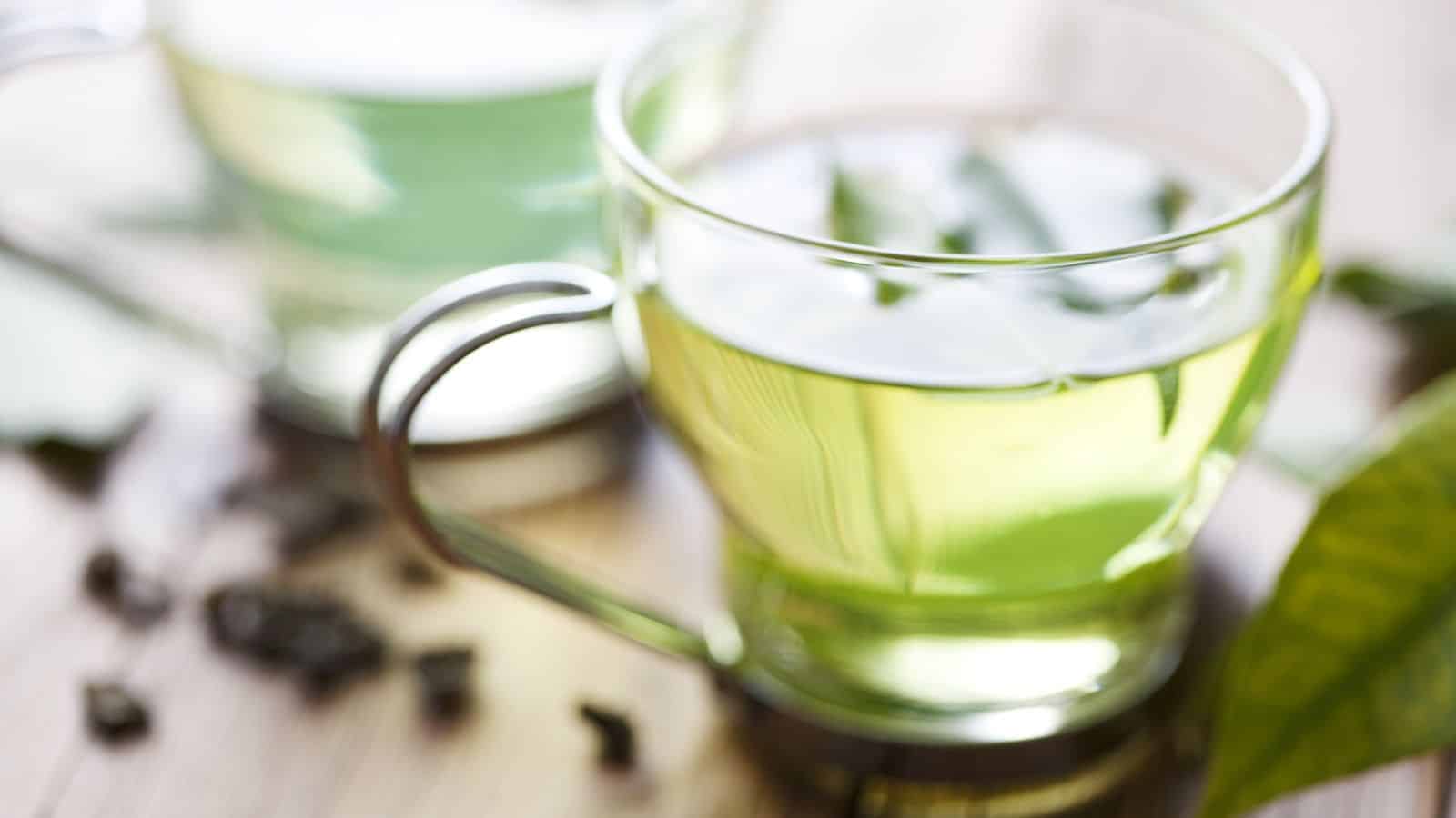 Two clear glass cups filled with green tea are placed on a wooden surface. One cup is in focus, showing light green liquid with leaves inside, while the other is blurred in the background. Loose tea leaves are scattered on the table.