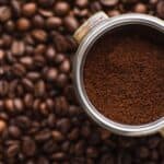 A metal portafilter filled with ground coffee is positioned over a surface covered with roasted coffee beans. The focus is on the rich, brown texture of the coffee grounds, contrasting with the shiny metal of the portafilter and the dark beans below.