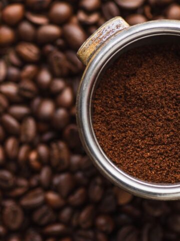 A metal portafilter filled with ground coffee is positioned over a surface covered with roasted coffee beans. The focus is on the rich, brown texture of the coffee grounds, contrasting with the shiny metal of the portafilter and the dark beans below.
