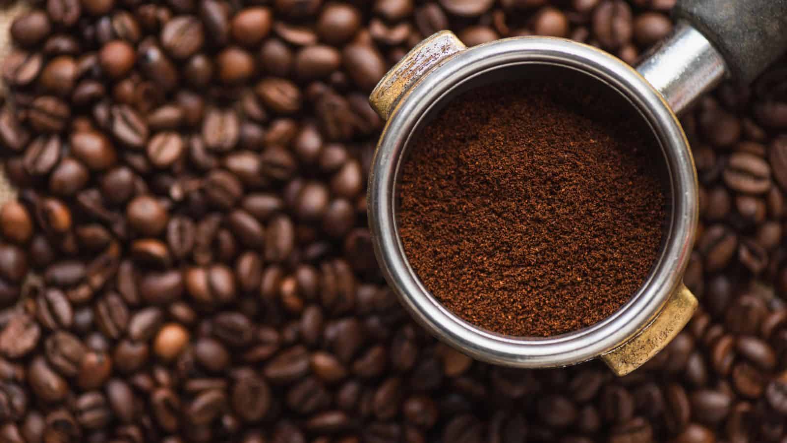 A metal portafilter filled with ground coffee is positioned over a surface covered with roasted coffee beans. The focus is on the rich, brown texture of the coffee grounds, contrasting with the shiny metal of the portafilter and the dark beans below.