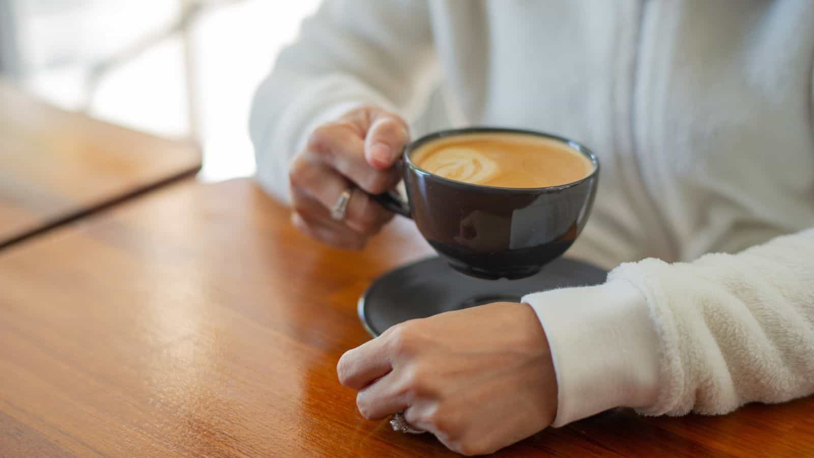 A person in a white sweater holds a black cup filled with coffee, seated at a wooden table. A saucer is placed beneath the cup. The background is softly blurred, suggesting a cozy setting, possibly a caf&eacute; or home.
