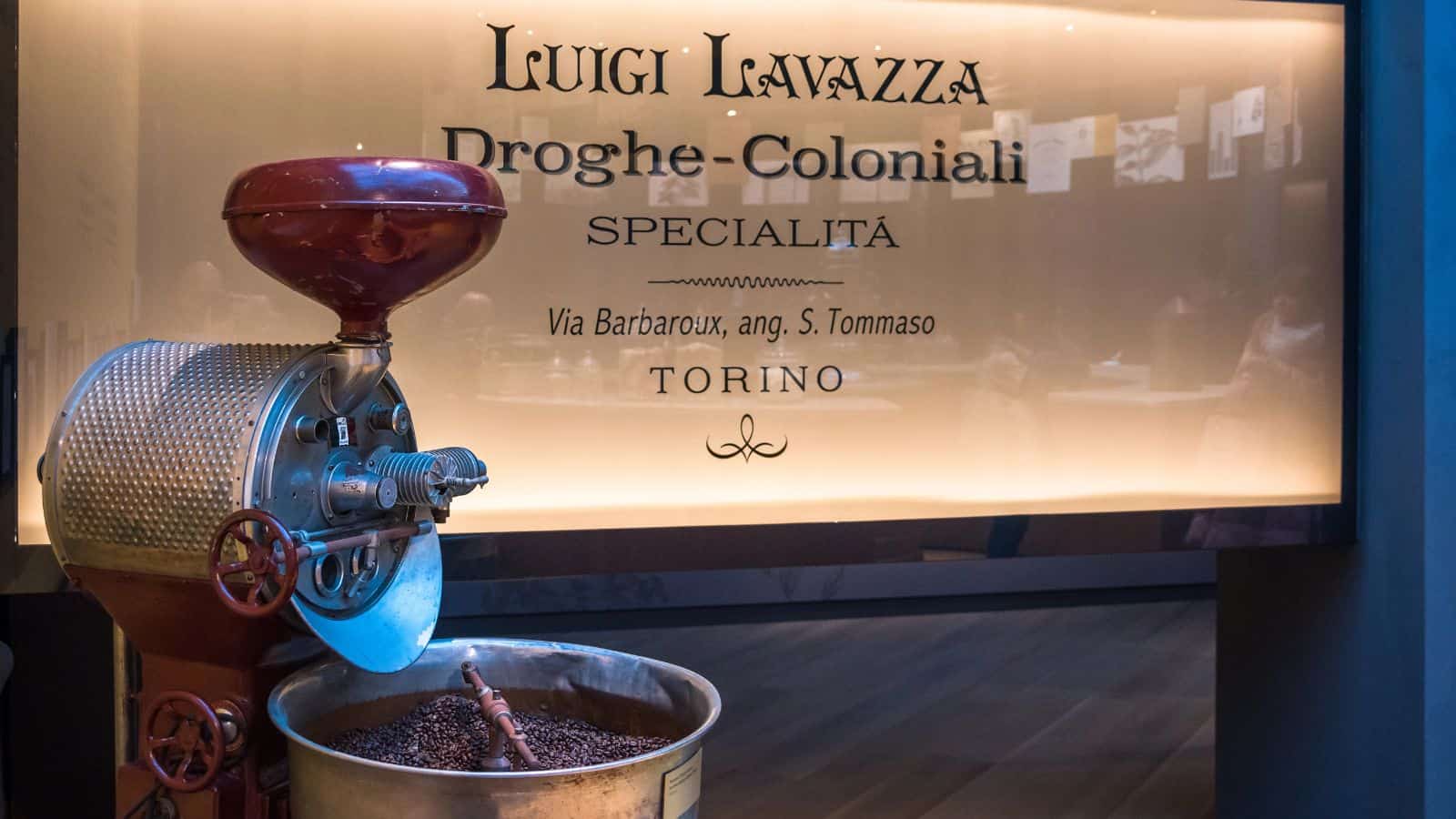A vintage coffee grinder sits on display with coffee beans in a metal container. The background shows a sign reading "Luigi Lavazza, Droghe-Coloniali, Specialit&agrave;, Via Barbaroux, ang. S. Tommaso, Torino" on a warmly lit wall.