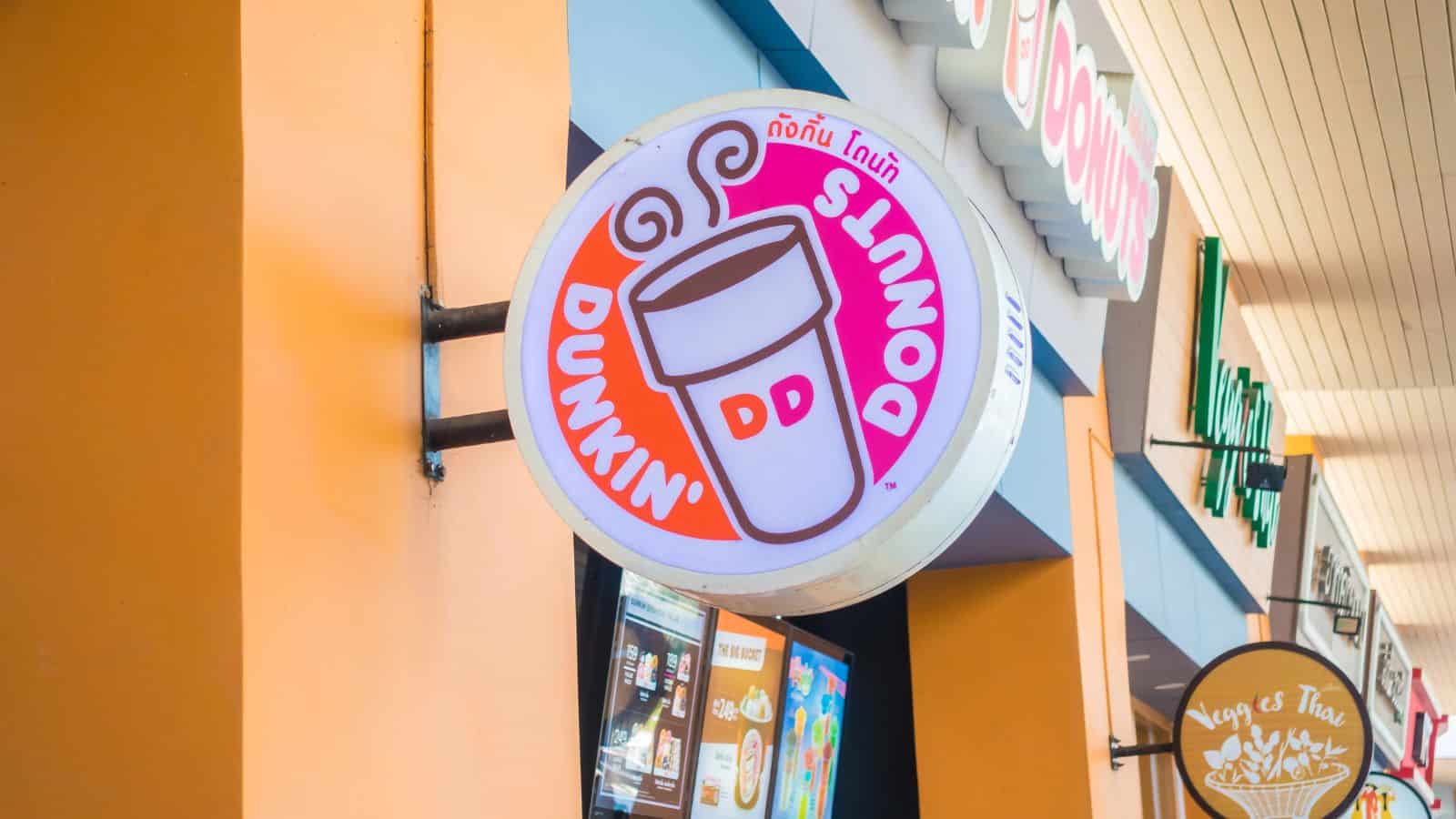 A Dunkin' Donuts sign featuring the brand's logo with a coffee cup and the text "Dunkin' Donuts" in English and Thai. The sign is attached to a storefront with other visible signs nearby.