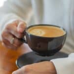 A person wearing a white sweater holds a black cup filled with latte. The cup rests on a matching saucer placed on a wooden surface. The person's other hand is on the table, showing no visible objects or markings.