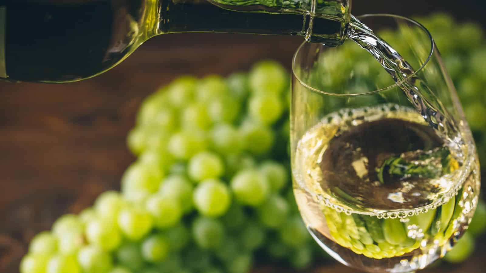 A bottle is pouring white wine into a glass, with a cluster of green grapes in the blurred background. The surface is wooden, adding a rustic atmosphere to the scene.