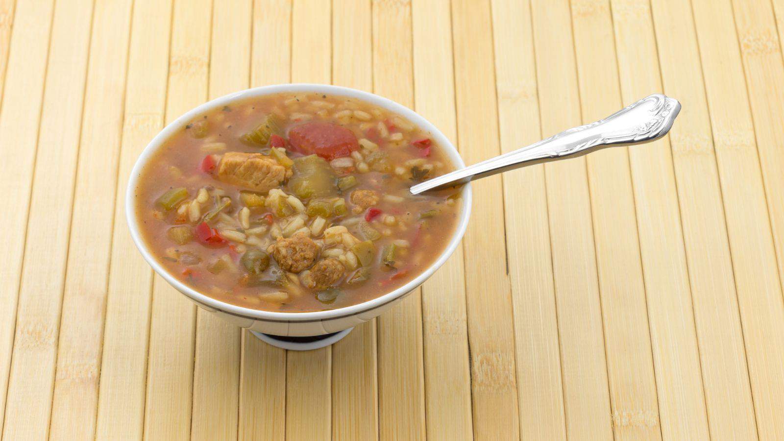 A bowl of soup containing chunks of meat, vegetables, and rice is placed on a bamboo surface. A silver spoon rests inside the bowl, and the soup appears thick and hearty with visible pieces of tomato and green vegetables.