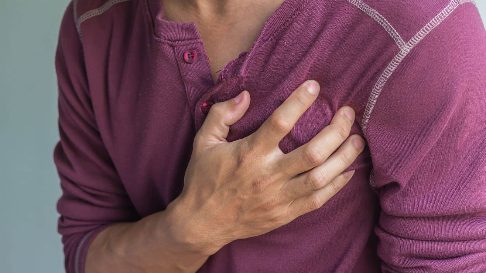 A person wearing a maroon long-sleeve shirt holds their hand to their chest, slightly tilting forward. The background is a plain, light color. The image conveys a sense of discomfort or pain in the chest area.