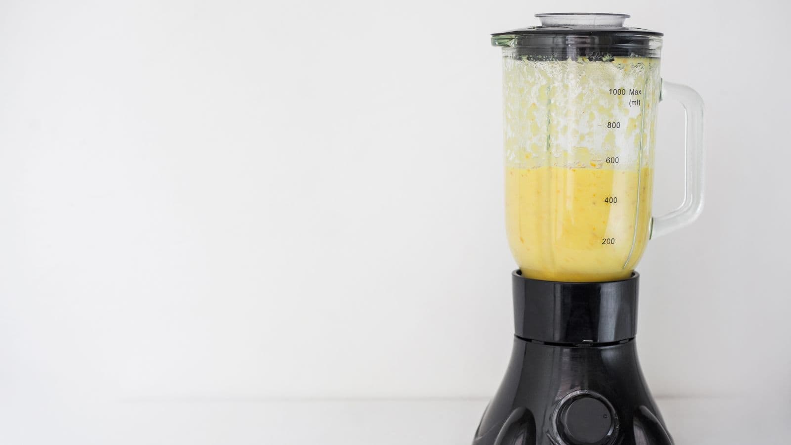 A black countertop blender with a transparent glass pitcher is filled with a yellow mixture. The pitcher has measurement markings. The background is plain white, creating a clean and minimalistic look.