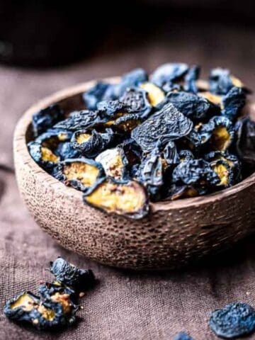 A bowl of dried black lemons sits on a dark fabric surface. Some pieces have spilled out of the bowl. The lemons are dark and wrinkled, with a textured appearance. The bowl is made of a rustic material.