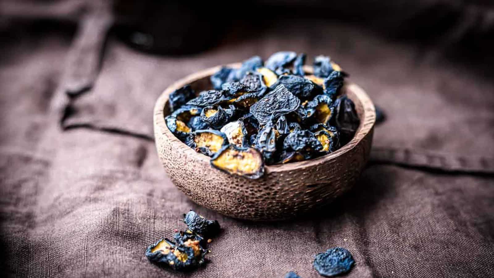 A bowl of dried black lemons sits on a dark fabric surface. Some pieces have spilled out of the bowl. The lemons are dark and wrinkled, with a textured appearance. The bowl is made of a rustic material.