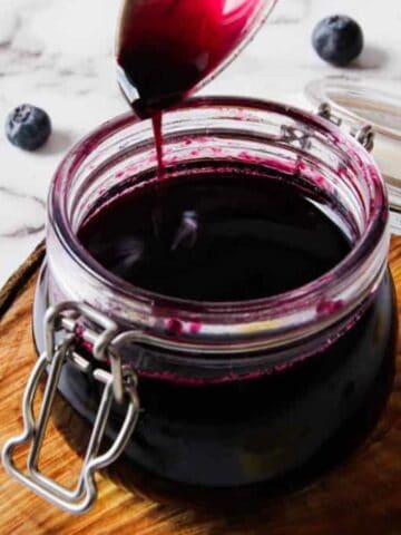 A jar of dark blueberry syrup sits on a wooden board. The lid is open as a spoon drizzles more syrup into the jar. Several blueberries are scattered around on a marble surface.