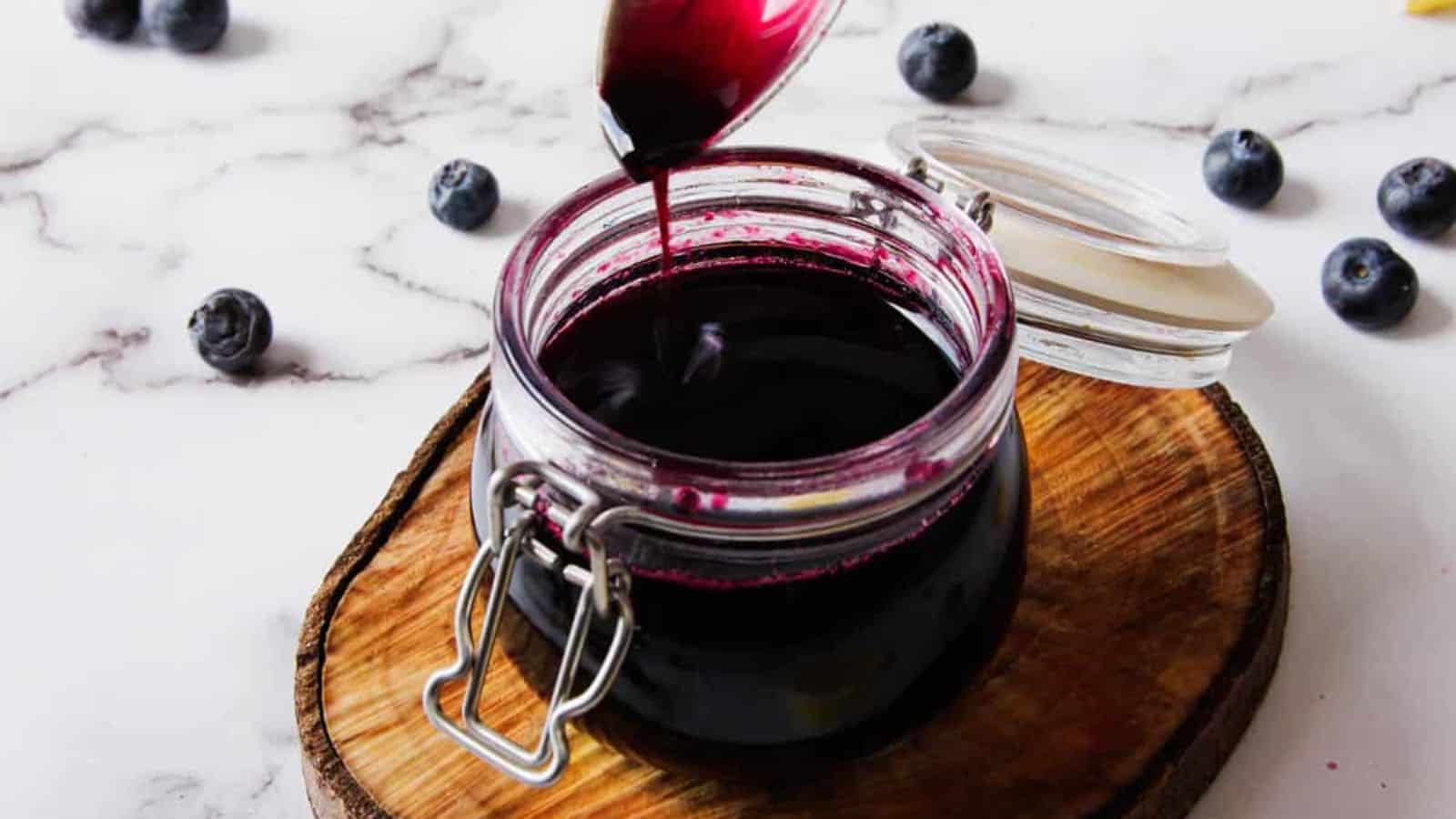 A jar of dark blueberry syrup sits on a wooden board. The lid is open as a spoon drizzles more syrup into the jar. Several blueberries are scattered around on a marble surface.