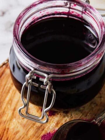 A jar of dark blueberry jam sits on a round wooden board with a silver spoon beside it. A few blueberries are scattered on the white marble surface in the background.