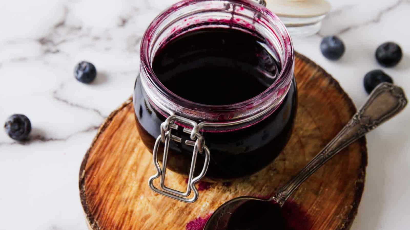 A jar of dark blueberry syrup sits on a wooden board. The lid is open as a spoon drizzles more syrup into the jar. Several blueberries are scattered around on a marble surface.