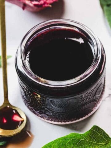 A jar filled with dark red liquid sits on a white surface surrounded by green leaves. A gold spoon with a small amount of the liquid is beside the jar. Partially visible pomegranate pieces are also scattered around.