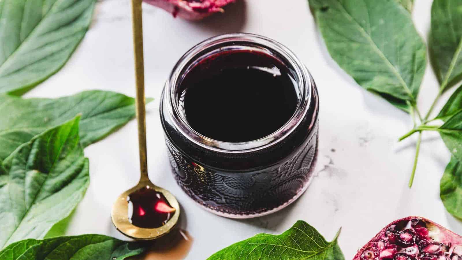A spoon with dark liquid is held above a glass jar filled with the same liquid. The jar is placed on a white surface, surrounded by green leaves. The liquid has a rich, deep color.