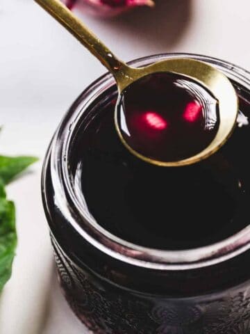 A spoon with dark liquid is held above a glass jar filled with the same liquid. The jar is placed on a white surface, surrounded by green leaves. The liquid has a rich, deep color.