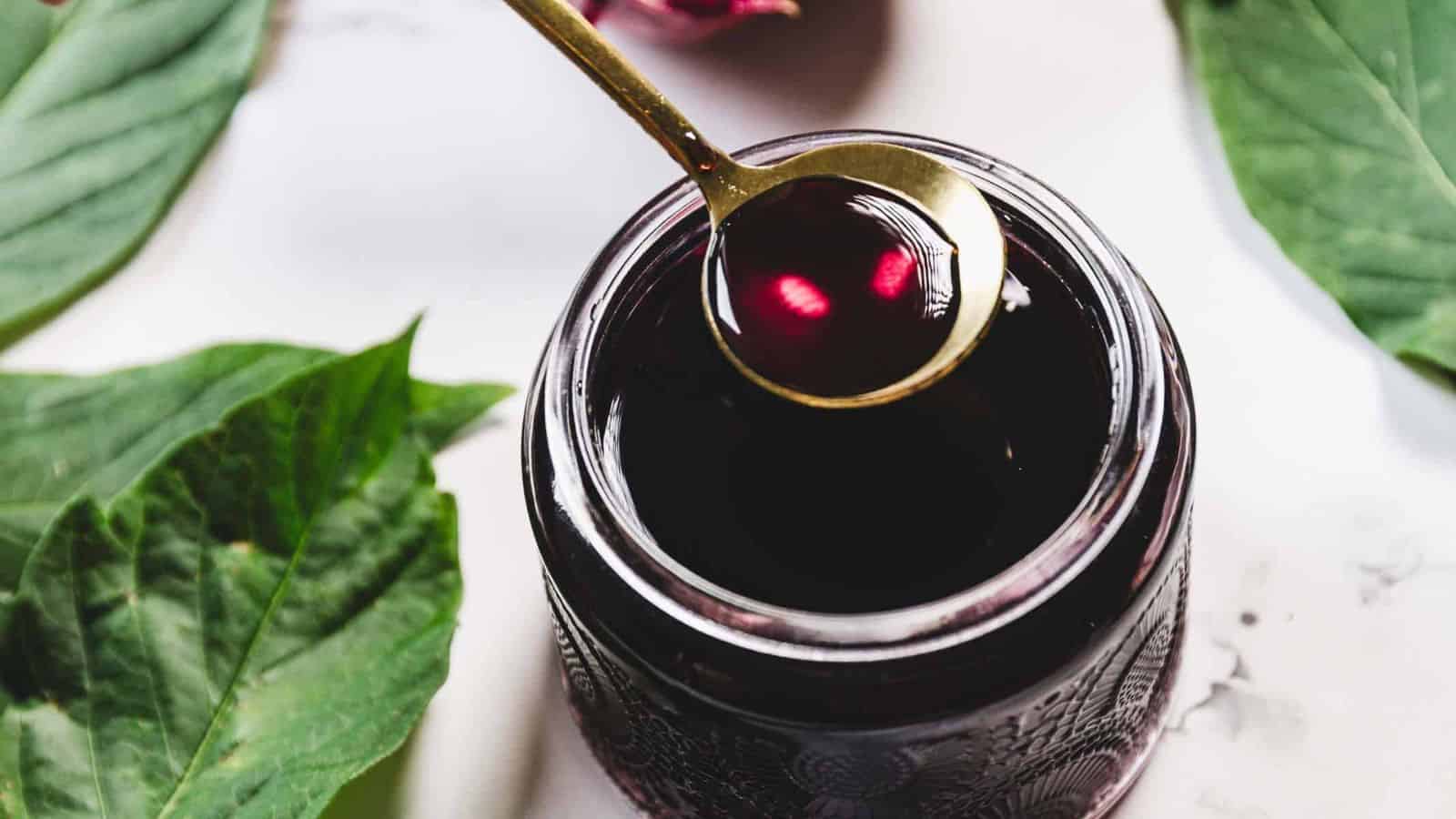 A spoon with dark liquid is held above a glass jar filled with the same liquid. The jar is placed on a white surface, surrounded by green leaves. The liquid has a rich, deep color.
