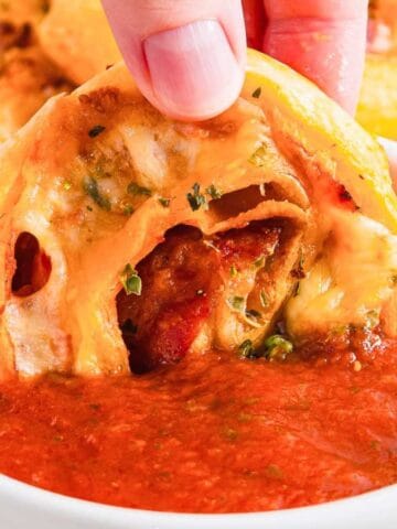 A hand dipping a cheesy, spiral-shaped pastry into a bowl of tomato sauce. The pastry appears crispy with herbs visible on the surface. A piece of basil is seen in the background on a wooden surface.