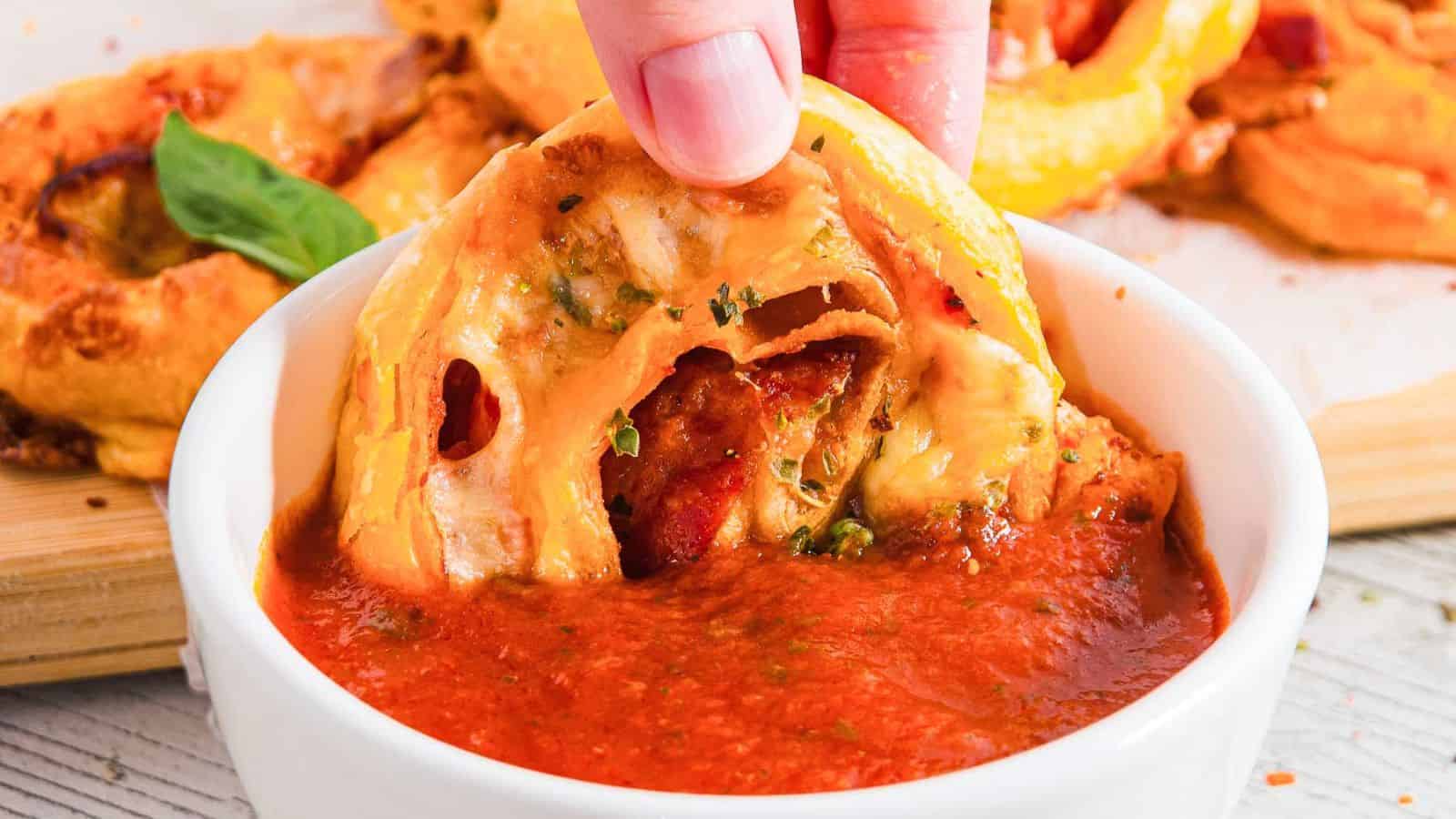 A hand dips a slice of pizza roll with visible cheese and pepperoni into a bowl of red marinara sauce. A basil leaf is partially visible in the background, resting on a wooden surface.