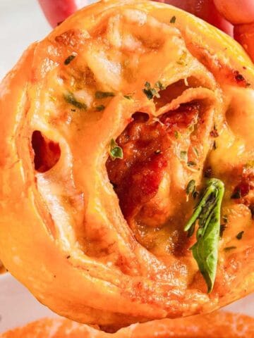 A hand holding a pizza roll. The roll is filled with melted cheese, tomato sauce, herbs, and pepperoni. Some basil leaves are visible. A bowl of dipping sauce is partially visible at the bottom of the image.