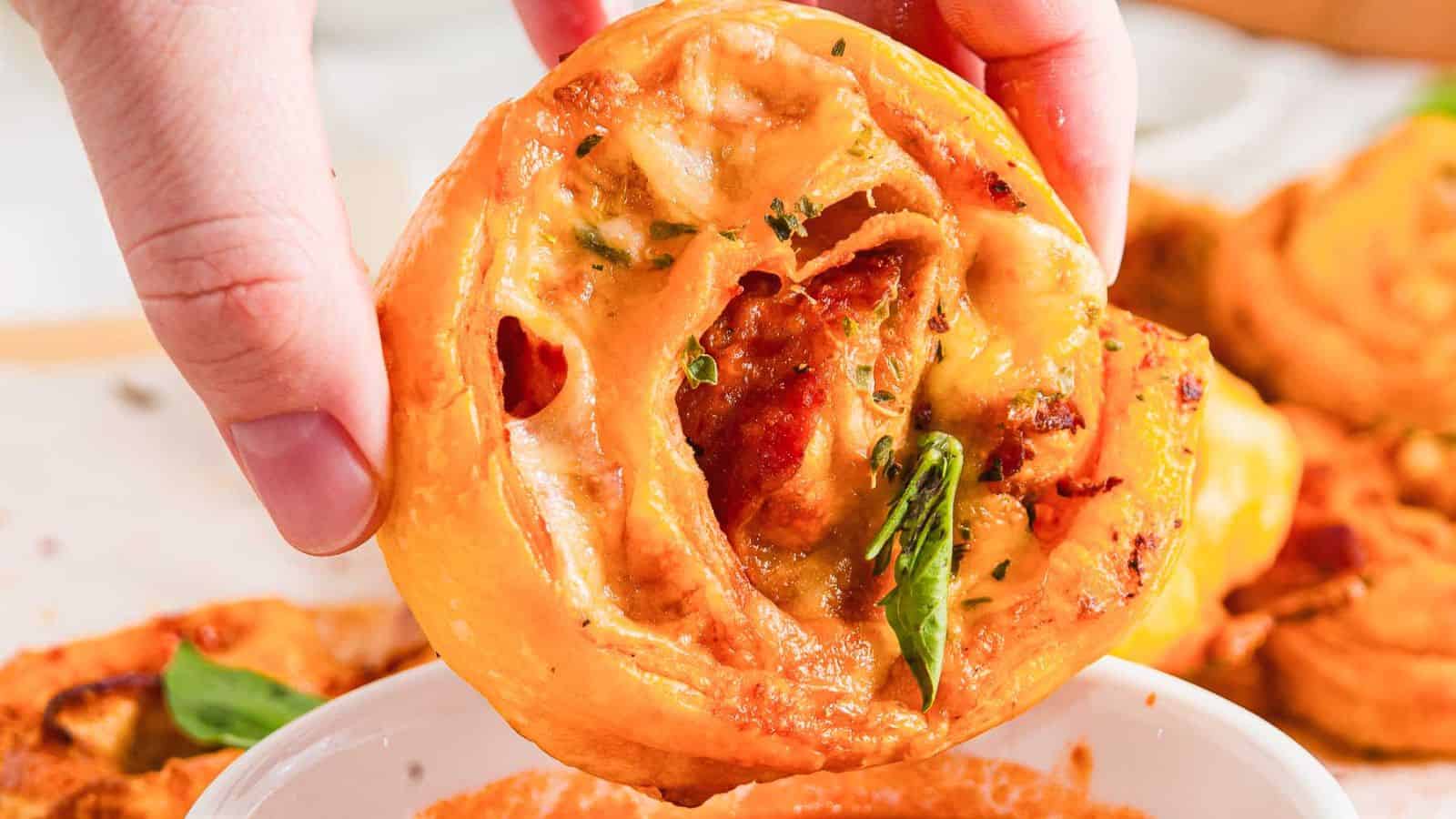 A hand holding a pizza roll. The roll is filled with melted cheese, tomato sauce, herbs, and pepperoni. Some basil leaves are visible. A bowl of dipping sauce is partially visible at the bottom of the image.