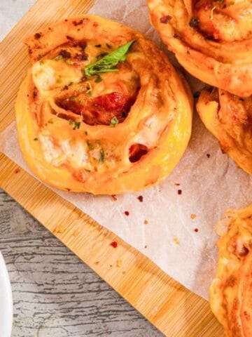 A wooden board with several spiral pizza rolls topped with basil leaves sits on a light surface. A small bowl of marinara sauce is placed to the side. The pizza rolls appear to have cheese and tomato filling.